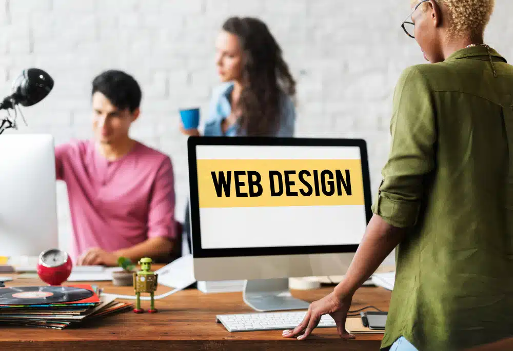 A diverse team working in an office setting on a web design project. In the foreground, a woman with short hair and glasses is standing and looking at a computer monitor displaying the words "WEB DESIGN." In the background, a man and a woman are discussing something, with the man sitting at a desk and the woman holding a blue coffee cup. The desk is cluttered with various items including vinyl records and small figurines.