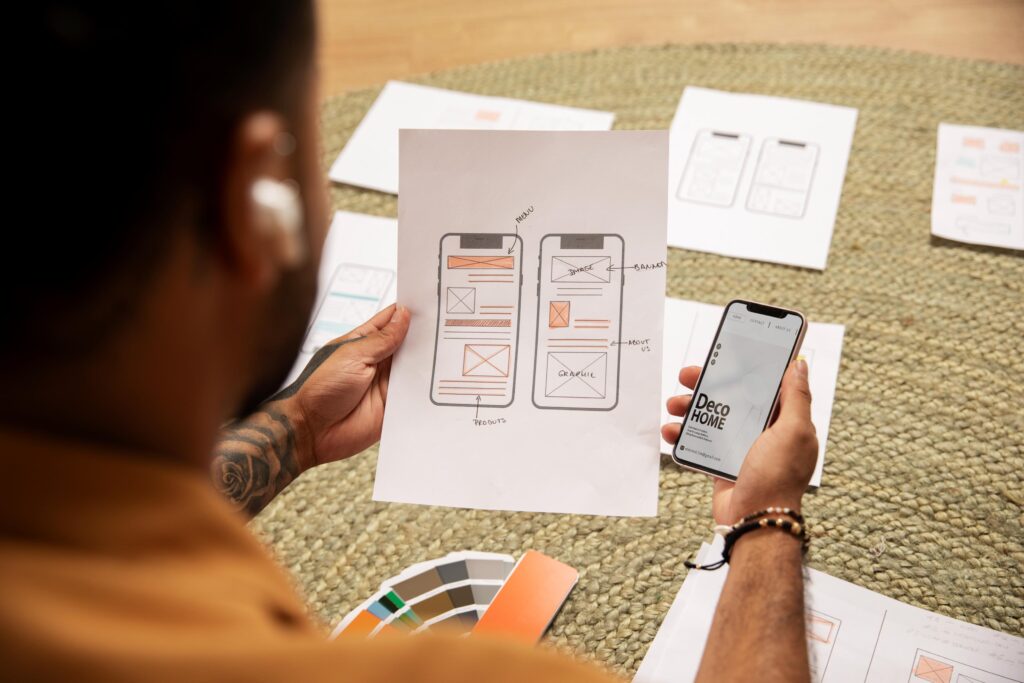 A person engaged in mobile app development, reviewing interface sketches and comparing them to a live design on a smartphone.
