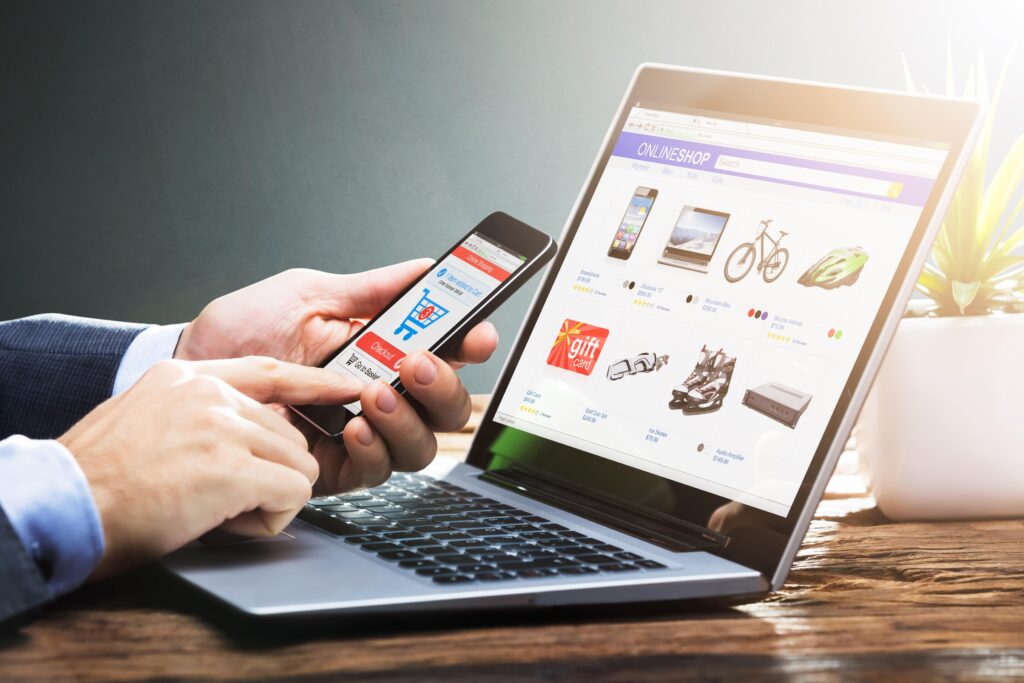 A person is using both a smartphone and a laptop to browse an online shop. The laptop screen displays a variety of products, including electronics, bicycles, and gift cards, while the smartphone shows a shopping cart page. The person's hands are interacting with the devices, emphasizing a multi-device online shopping experience. A potted plant is visible in the background, adding a touch of greenery to the workspace.