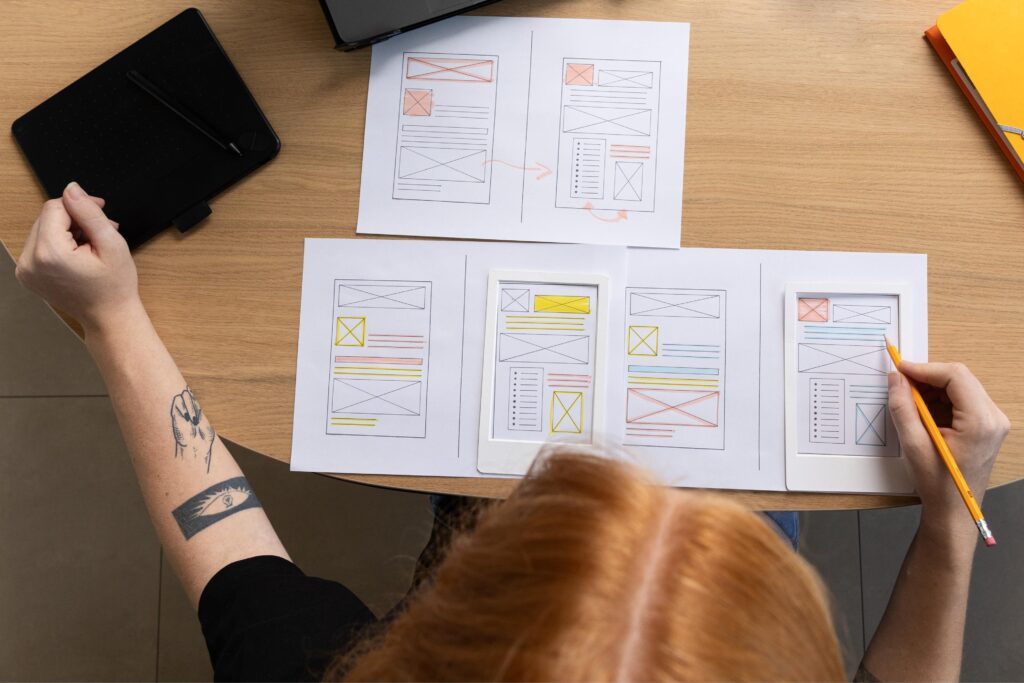 A person with red hair and a tattoo on their arm is working on website wireframes at a wooden desk. They are using a pencil to sketch out design ideas on multiple sheets of paper, which include colorful, annotated wireframe layouts. A black tablet with a stylus and a laptop are also on the desk, suggesting a digital design process.